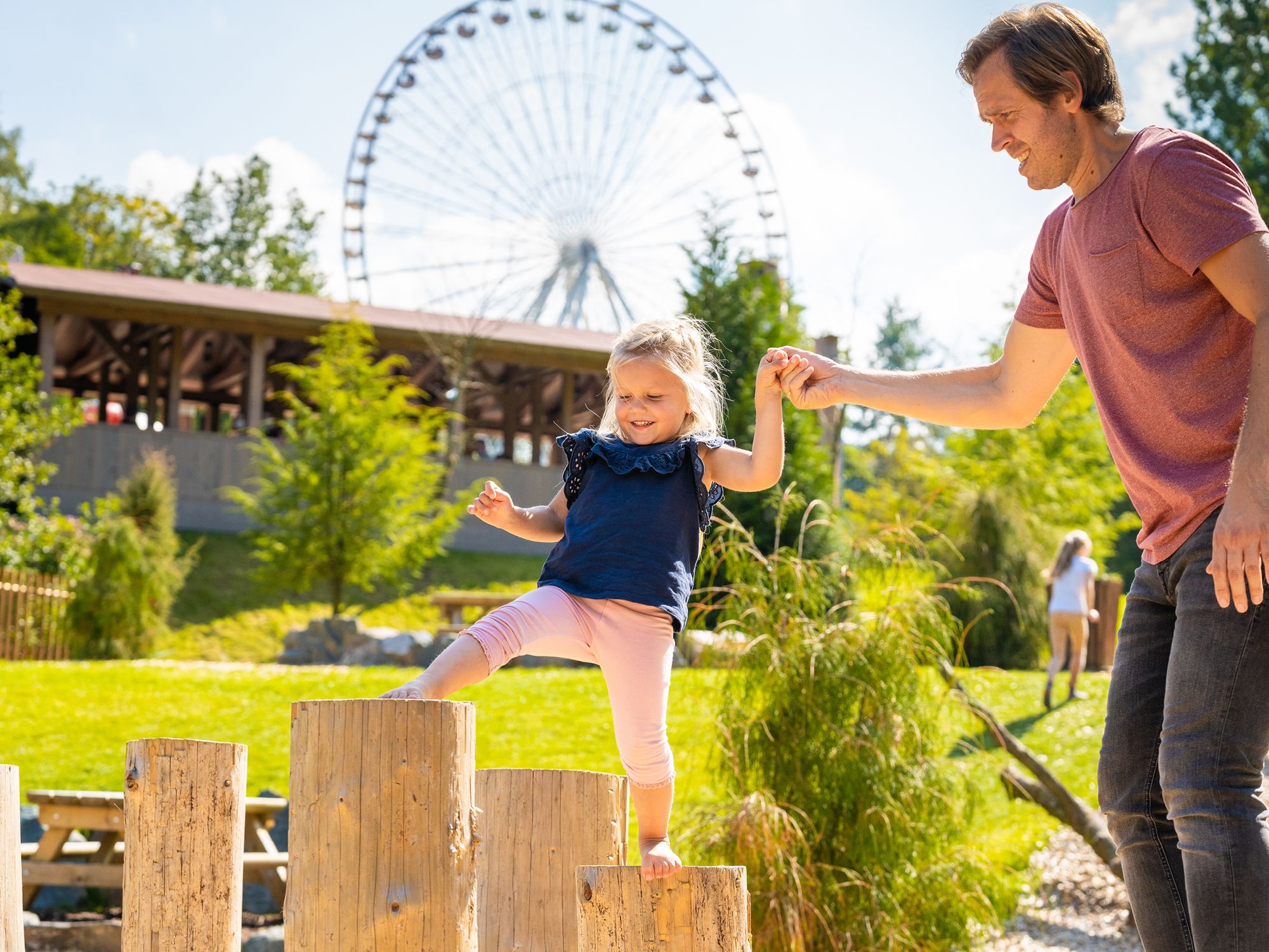 In der Adventure Area mit dem Riesenrad im Hintergrund probiert ein kleines Mädchen auf Baumstämmen zu laufen und der Papa hilft ihr dabei.